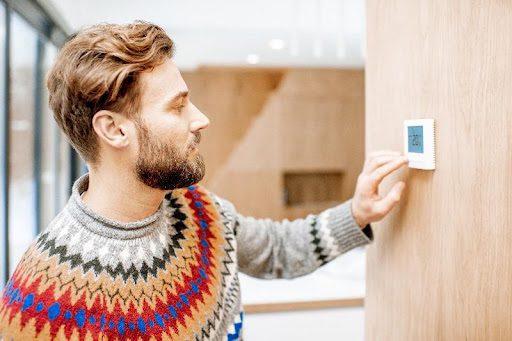 A man in a seasonal sweater adjusting the heat on his wall thermostat in Battlefield, MO. A homeowner increasing the heat of his furnace after a repair.