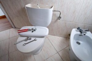 Bathroom with tools on a toilet, including wrenches and pliers. A bidet is adjacent to the toilet. The toilet tank lid is removed.