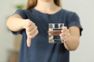 Person in a blue shirt holding a glass of water with one hand and showing a thumbs-down with the other hand.