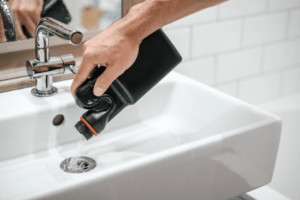 A hand pouring liquid from a black bottle into a white sink.