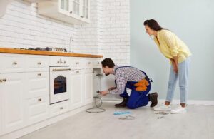 A technician in blue overalls works under a kitchen cabinet while a woman in a yellow shirt observes. Tools are scattered on the floor.