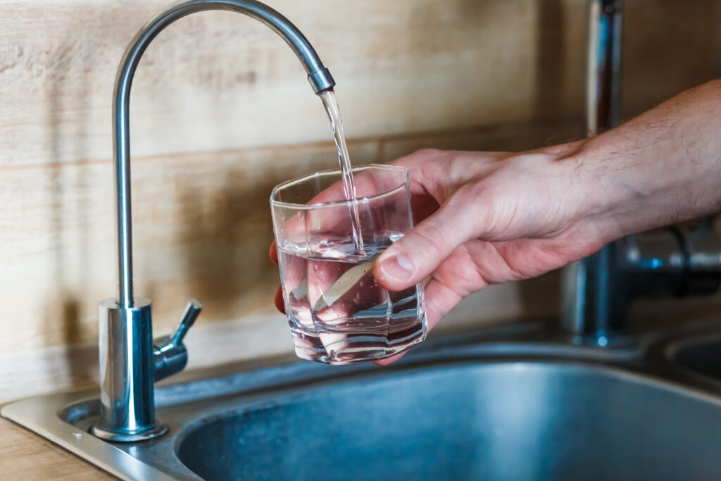 A hand fills a clear glass with water from a kitchen faucet above a stainless steel sink.
