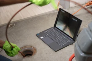 Person wearing green gloves inserts a cable into a drain while viewing footage on a nearby laptop screen.